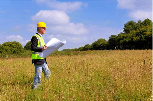 A person wearing construction jacket looking at big paper with plans standing on a grass field.
